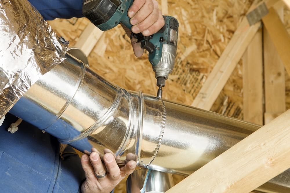 Worker drills a screw into a shiny metal HVAC duct in an unfinished attic.
